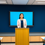 A professional female early childhood education instructor. She is wearing modest business casual attire and professional dress. She stands confidently at a modern podium in a bright lecture hall, engaging with an unseen adult audience. A large screen behind her displays educational content related to child development and modern learning methods, such as graphs or child-friendly illustrations. The atmosphere is inspiring and educational, with soft, professional lighting. This is a high-resolution, professional photograph with sharp focus. fully clothed, modest clothing, appropriate attire, professional dress, safe for work, appropriate content, perfect anatomy, correct proportions, natural pose, well-formed hands, proper finger count, natural body proportions.